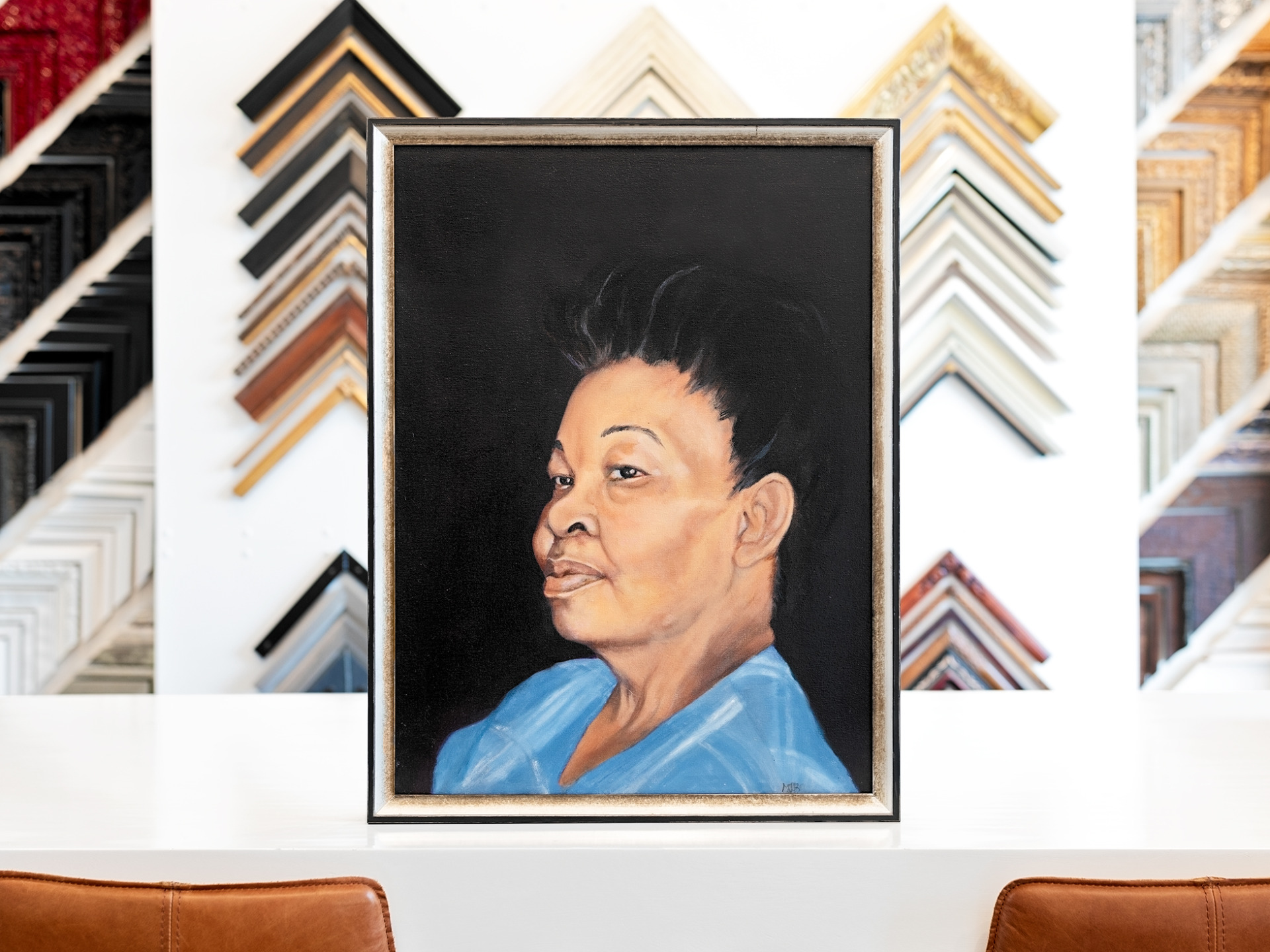 Portrait painting of an older woman, displayed on a white table in a thin gold, silver and black edged frame.